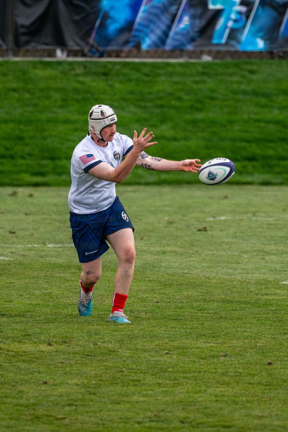 A rugby player wearing a white jersey with a US flag patch, navy blue shorts, and a white Canterbury (CCC) headguard is in the act of passing a rugby ball on a grass pitch. The player appears to be executing a lateral or spin pass, with the ball just leaving or about to be caught by their hands. Background shows a grassy embankment and blue advertising banners.