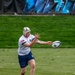 A rugby player wearing a white jersey with a US flag patch, navy blue shorts, and a white Canterbury (CCC) headguard is in the act of passing a rugby ball on a grass pitch. The player appears to be executing a lateral or spin pass, with the ball just leaving or about to be caught by their hands. Background shows a grassy embankment and blue advertising banners.
