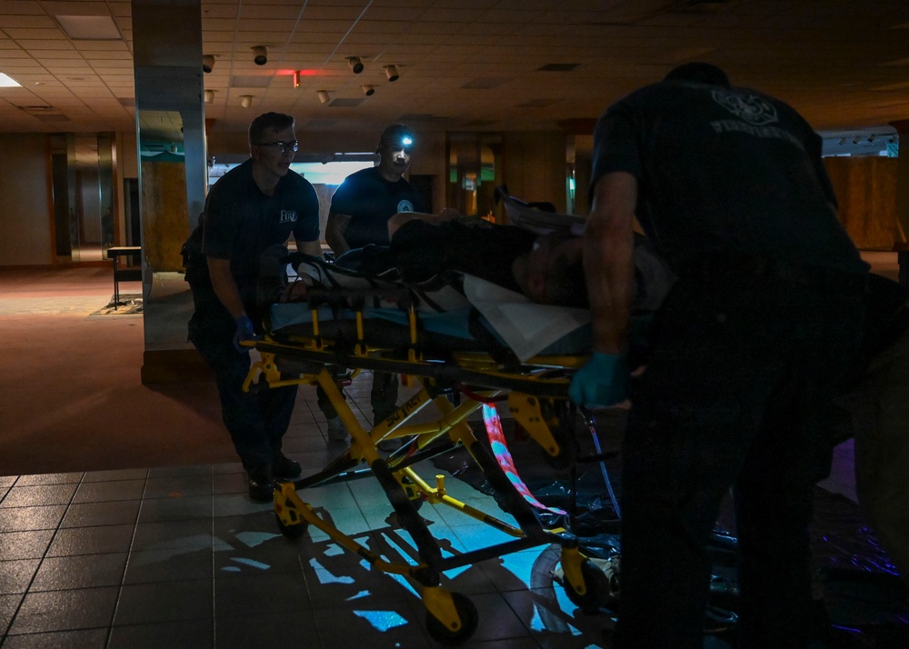 Emergency medical personnel are transporting a patient on a yellow wheeled stretcher through a darkened indoor corridor. At least three responders wearing dark uniforms and blue gloves are visible, with one using a headlamp to illuminate the scene. The setting appears to be a large building interior with low ambient lighting.