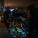 Emergency medical personnel are transporting a patient on a yellow wheeled stretcher through a darkened indoor corridor. At least three responders wearing dark uniforms and blue gloves are visible, with one using a headlamp to illuminate the scene. The setting appears to be a large building interior with low ambient lighting.