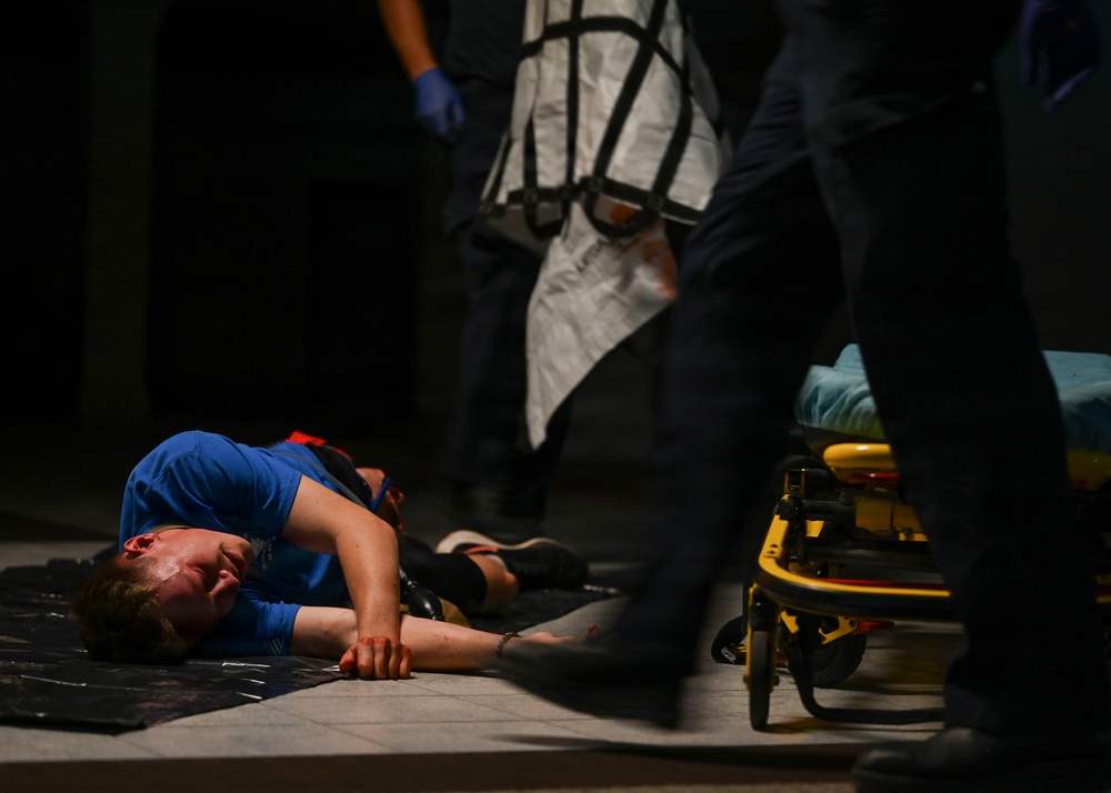 A person in a blue shirt is lying on the ground, apparently injured or incapacitated. Emergency medical personnel are present, visible by blue medical gloves and a yellow gurney/stretcher nearby. The scene appears to be an active medical emergency response.