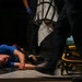A person in a blue shirt is lying on the ground, apparently injured or incapacitated. Emergency medical personnel are present, visible by blue medical gloves and a yellow gurney/stretcher nearby. The scene appears to be an active medical emergency response.