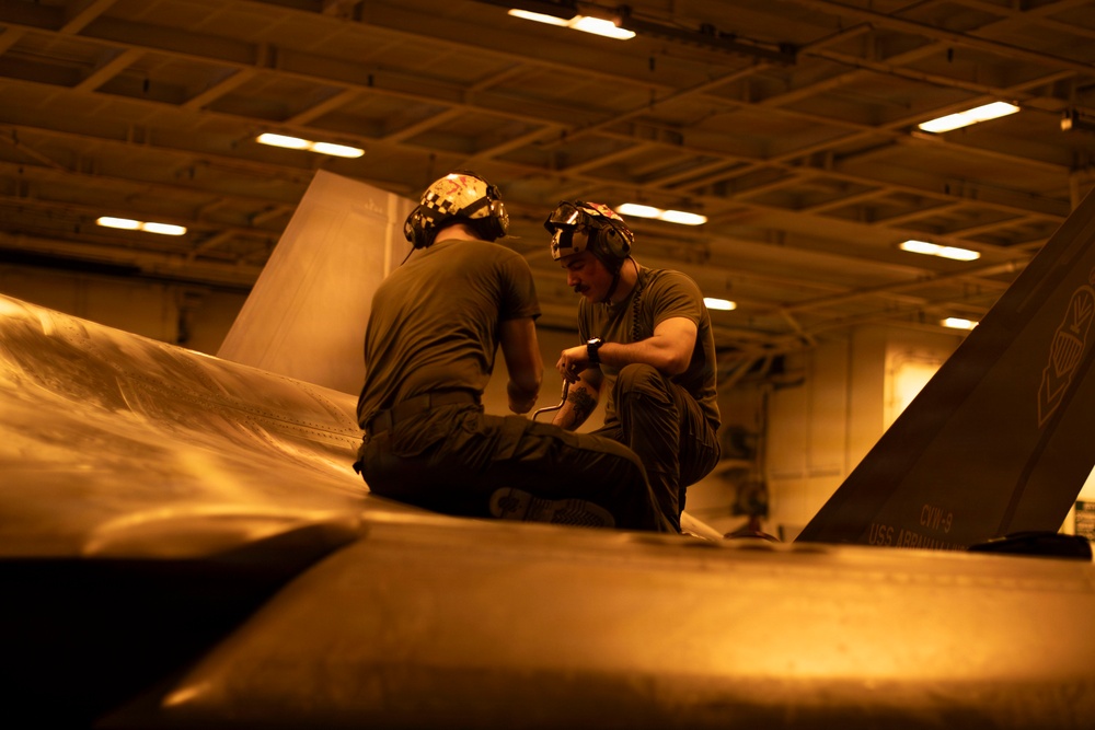 Two military aviation maintenance personnel are crouched on top of a naval aircraft inside what appears to be a carrier hangar bay, performing maintenance or inspection work. The scene is bathed in warm amber/orange lighting typical of a ship's hangar deck environment. Text visible on the aircraft's tail fin references 'CVW-9' and 'USS' indicating this is aboard a U.S. Navy aircraft carrier.