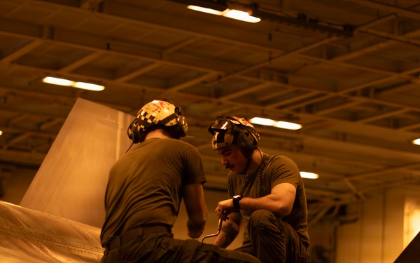 Two military aviation maintenance personnel are crouched on top of a naval aircraft inside what appears to be a carrier hangar bay, performing maintenance or inspection work. The scene is bathed in warm amber/orange lighting typical of a ship's hangar deck environment. Text visible on the aircraft's tail fin references 'CVW-9' and 'USS' indicating this is aboard a U.S. Navy aircraft carrier.