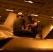 Two military aviation maintenance personnel are crouched on top of a naval aircraft inside what appears to be a carrier hangar bay, performing maintenance or inspection work. The scene is bathed in warm amber/orange lighting typical of a ship's hangar deck environment. Text visible on the aircraft's tail fin references 'CVW-9' and 'USS' indicating this is aboard a U.S. Navy aircraft carrier.