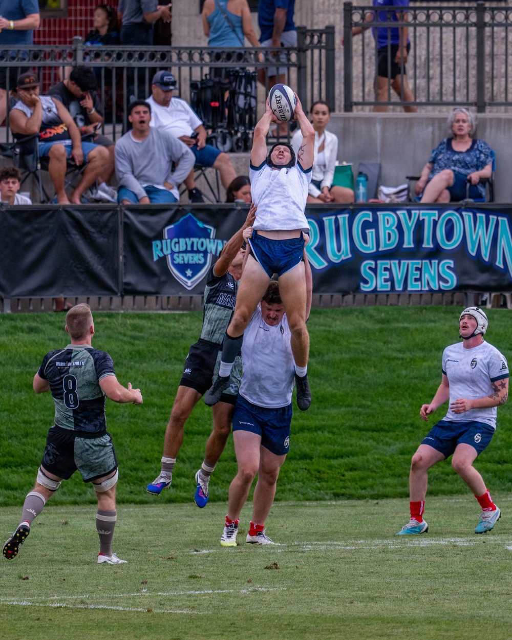 A rugby sevens line-out is being contested during a match at the 'Rugbytown Sevens' tournament. A player in a white and blue kit is being lifted by a teammate to catch a Gilbert rugby ball at height, while opposing players in dark camouflage-style uniforms attempt to contest. Spectators are visible in the background along with prominent tournament branding banners.