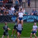 A rugby sevens line-out is being contested during a match at the 'Rugbytown Sevens' tournament. A player in a white and blue kit is being lifted by a teammate to catch a Gilbert rugby ball at height, while opposing players in dark camouflage-style uniforms attempt to contest. Spectators are visible in the background along with prominent tournament branding banners.