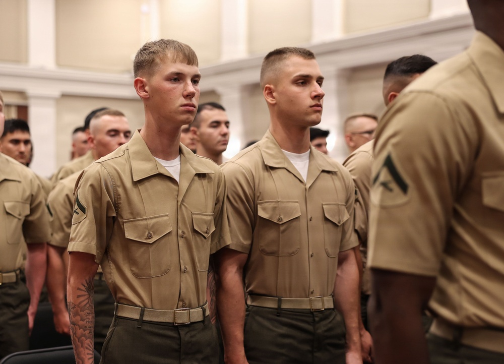 A group of U.S. Marine Corps personnel standing in formation in what appears to be an indoor ceremonial or administrative setting. The individuals are wearing the standard khaki/tan Service Charlie uniform with olive drab trousers. The two foreground Marines display Private First Class rank insignia on their collars.