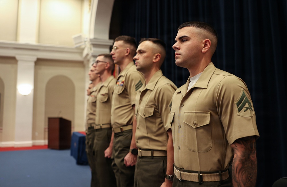 Four U.S. Marine Corps personnel are standing at attention in a formal indoor setting, likely during a ceremony or official event. They are wearing the standard khaki service uniform (Charlie uniform) with green trousers. The Marines are arranged in a diagonal line, facing the same direction in a disciplined formation.