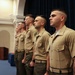 Four U.S. Marine Corps personnel are standing at attention in a formal indoor setting, likely during a ceremony or official event. They are wearing the standard khaki service uniform (Charlie uniform) with green trousers. The Marines are arranged in a diagonal line, facing the same direction in a disciplined formation.