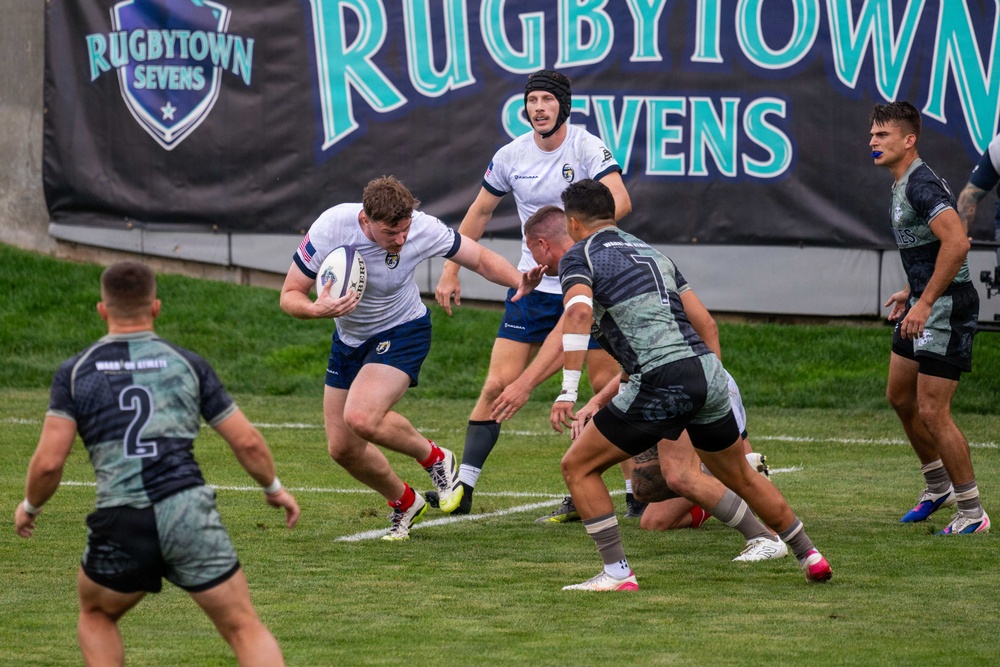 A rugby sevens match is in progress at the Rugbytown Sevens tournament, with players in white uniforms (appearing to represent the USA based on the flag patch) competing against players in dark camouflage-patterned jerseys. The ball carrier in white is attempting to evade two defenders while a teammate in a scrum cap provides support from behind. The action takes place on a well-maintained grass field in front of prominent tournament branding banners.
