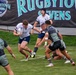 A rugby sevens match is in progress at the Rugbytown Sevens tournament, with players in white uniforms (appearing to represent the USA based on the flag patch) competing against players in dark camouflage-patterned jerseys. The ball carrier in white is attempting to evade two defenders while a teammate in a scrum cap provides support from behind. The action takes place on a well-maintained grass field in front of prominent tournament branding banners.
