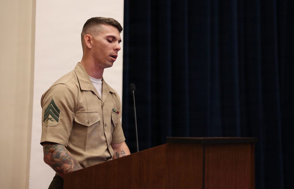 A U.S. Marine Corps Sergeant stands at a wooden podium, appearing to speak or read from notes. He is wearing the tan/khaki service uniform with visible rank insignia (Sergeant chevrons) on his left sleeve and a small ribbon rack on his chest. His arms display extensive tattoos, and a microphone is positioned in front of him against a backdrop of dark blue curtains.