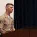 A U.S. Marine Corps Sergeant stands at a wooden podium, appearing to speak or read from notes. He is wearing the tan/khaki service uniform with visible rank insignia (Sergeant chevrons) on his left sleeve and a small ribbon rack on his chest. His arms display extensive tattoos, and a microphone is positioned in front of him against a backdrop of dark blue curtains.
