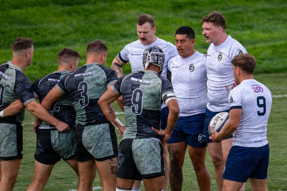 A rugby match or pre-match interaction is taking place between two teams on a green grass field. One team wears camouflage-patterned gray/black jerseys labeled 'Warrior Athlete' with visible numbers 3 and 9, while the opposing team wears white jerseys with blue shorts branded 'Jakuma' and featuring an 'America 250' logo. Players from both teams appear to be in a post-play or pre-set discussion near the center of the field.