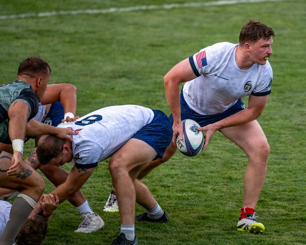 A rugby match is in progress on a well-maintained grass field. A player wearing a white and blue Akuma jersey with a US flag patch on the sleeve is carrying the ball and breaking away from a ruck or maul. Opposing players in camouflage-patterned uniforms are engaged in the tackle/breakdown area.