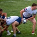 A rugby match is in progress on a well-maintained grass field. A player wearing a white and blue Akuma jersey with a US flag patch on the sleeve is carrying the ball and breaking away from a ruck or maul. Opposing players in camouflage-patterned uniforms are engaged in the tackle/breakdown area.