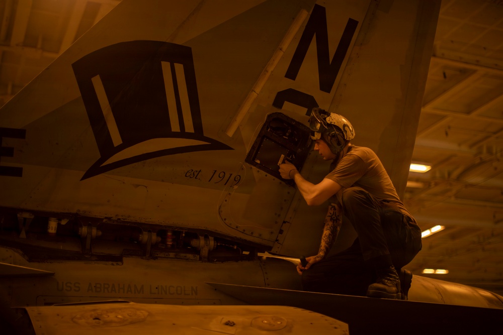 A military aircraft maintainer or technician is performing maintenance work on a naval aircraft aboard USS Abraham Lincoln, working on what appears to be an avionics or equipment bay access panel. The aircraft bears a squadron insignia depicting a top hat with the text 'est. 1919' and the tail code letter 'N'. The image is taken in a ship's hangar bay under warm amber lighting conditions.
