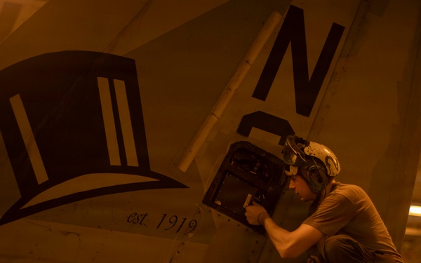 A military aircraft maintainer or technician is performing maintenance work on a naval aircraft aboard USS Abraham Lincoln, working on what appears to be an avionics or equipment bay access panel. The aircraft bears a squadron insignia depicting a top hat with the text 'est. 1919' and the tail code letter 'N'. The image is taken in a ship's hangar bay under warm amber lighting conditions.