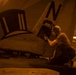 A military aircraft maintainer or technician is performing maintenance work on a naval aircraft aboard USS Abraham Lincoln, working on what appears to be an avionics or equipment bay access panel. The aircraft bears a squadron insignia depicting a top hat with the text 'est. 1919' and the tail code letter 'N'. The image is taken in a ship's hangar bay under warm amber lighting conditions.
