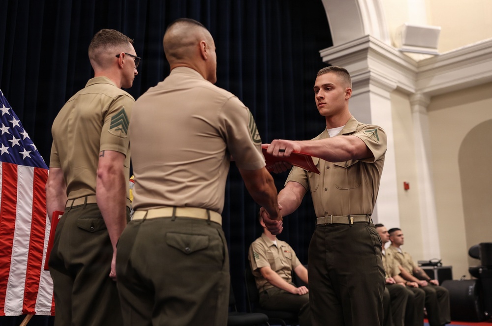 A formal military ceremony is taking place in an indoor venue, likely a ceremonial hall. A Marine is receiving what appears to be a folded document or certificate from a higher-ranking officer while another NCO stands nearby. Additional uniformed Marines are seated in the background as observers.