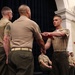 A formal military ceremony is taking place in an indoor venue, likely a ceremonial hall. A Marine is receiving what appears to be a folded document or certificate from a higher-ranking officer while another NCO stands nearby. Additional uniformed Marines are seated in the background as observers.