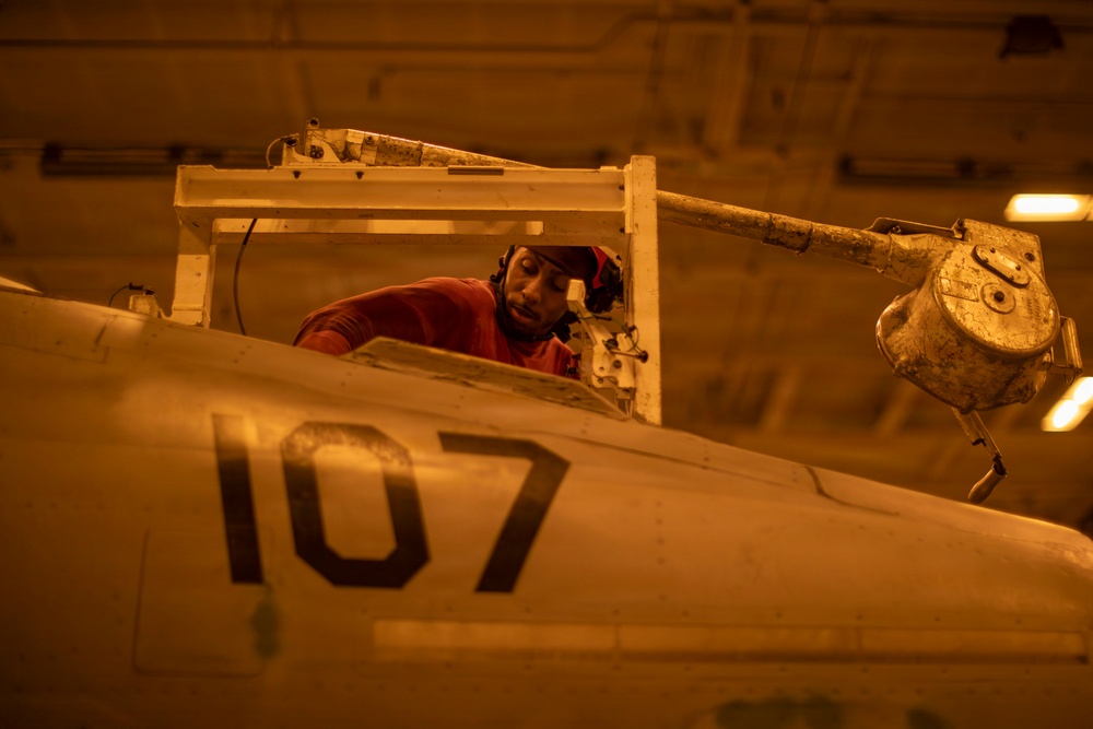 A technician or maintenance crew member wearing a red shirt and headgear is working inside the cockpit of a military aircraft marked with the number '107'. The work is being conducted in what appears to be a hangar bay, likely aboard a naval vessel, under warm amber/orange lighting. The scene depicts routine aircraft maintenance or inspection operations.