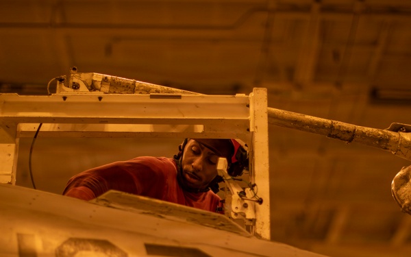 A technician or maintenance crew member wearing a red shirt and headgear is working inside the cockpit of a military aircraft marked with the number '107'. The work is being conducted in what appears to be a hangar bay, likely aboard a naval vessel, under warm amber/orange lighting. The scene depicts routine aircraft maintenance or inspection operations.