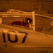 A technician or maintenance crew member wearing a red shirt and headgear is working inside the cockpit of a military aircraft marked with the number '107'. The work is being conducted in what appears to be a hangar bay, likely aboard a naval vessel, under warm amber/orange lighting. The scene depicts routine aircraft maintenance or inspection operations.