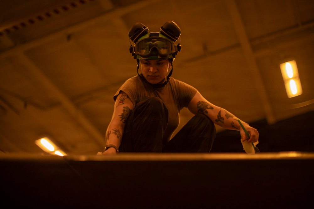 A person wearing a helmet with goggles pushed up on their forehead is crouched down and working on a surface in a large, dimly lit industrial or hangar-like space. The individual has visible tattoos on both arms and is wearing a brown t-shirt and dark pants. The environment is bathed in a warm orange/amber light, suggesting an aircraft hangar or similar military/industrial facility.