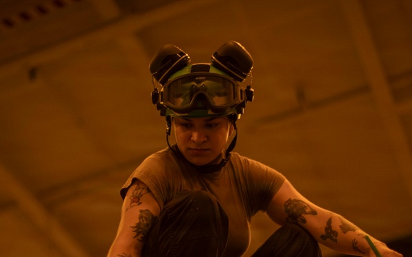 A person wearing a helmet with goggles pushed up on their forehead is crouched down and working on a surface in a large, dimly lit industrial or hangar-like space. The individual has visible tattoos on both arms and is wearing a brown t-shirt and dark pants. The environment is bathed in a warm orange/amber light, suggesting an aircraft hangar or similar military/industrial facility.