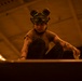 A person wearing a helmet with goggles pushed up on their forehead is crouched down and working on a surface in a large, dimly lit industrial or hangar-like space. The individual has visible tattoos on both arms and is wearing a brown t-shirt and dark pants. The environment is bathed in a warm orange/amber light, suggesting an aircraft hangar or similar military/industrial facility.