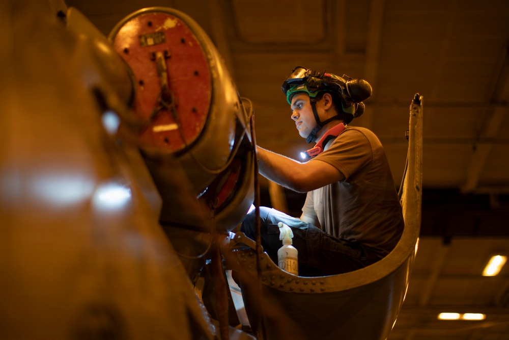 A technician or mechanic is performing an inspection or maintenance task on what appears to be a large aircraft engine or propeller assembly in a hangar or maintenance facility. The worker is equipped with a helmet-mounted headlamp and goggles, using a handheld light source to illuminate the work area. The warm amber lighting of the facility contrasts with the bright white light from the worker's equipment.