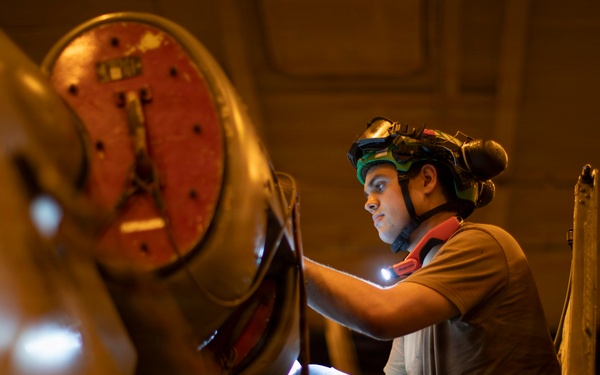 A technician or mechanic is performing an inspection or maintenance task on what appears to be a large aircraft engine or propeller assembly in a hangar or maintenance facility. The worker is equipped with a helmet-mounted headlamp and goggles, using a handheld light source to illuminate the work area. The warm amber lighting of the facility contrasts with the bright white light from the worker's equipment.