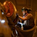 A technician or mechanic is performing an inspection or maintenance task on what appears to be a large aircraft engine or propeller assembly in a hangar or maintenance facility. The worker is equipped with a helmet-mounted headlamp and goggles, using a handheld light source to illuminate the work area. The warm amber lighting of the facility contrasts with the bright white light from the worker's equipment.