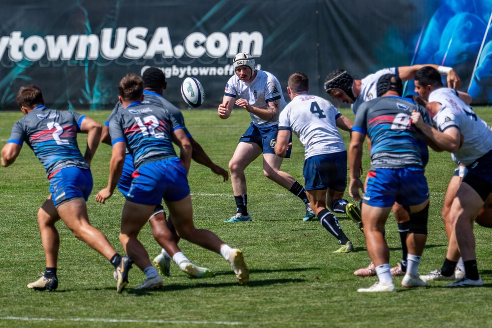 A rugby match is in progress on a well-maintained grass field, with two teams competing — one in white uniforms and one in blue/grey uniforms. A player wearing a white jersey and protective headgear is in the act of passing or kicking the ball. A banner in the background reads 'rugbytownUSA.com', suggesting this is an organized rugby event in the United States.