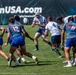 A rugby match is in progress on a well-maintained grass field, with two teams competing — one in white uniforms and one in blue/grey uniforms. A player wearing a white jersey and protective headgear is in the act of passing or kicking the ball. A banner in the background reads 'rugbytownUSA.com', suggesting this is an organized rugby event in the United States.