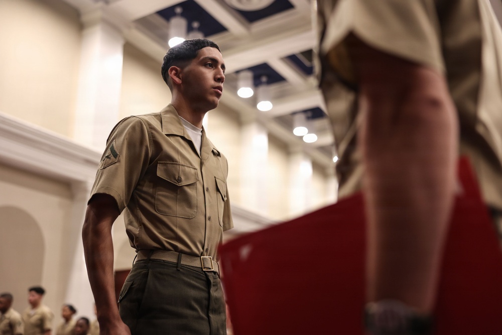A young male service member in U.S. Marine Corps Service Charlie uniform stands at attention or in formation inside a formal indoor venue. The setting appears to be a military ceremony or official event, indicated by the decorated ceiling and presence of multiple uniformed personnel. A second uniformed individual in the foreground holds a red folder or binder, partially obscuring the view.
