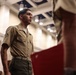 A young male service member in U.S. Marine Corps Service Charlie uniform stands at attention or in formation inside a formal indoor venue. The setting appears to be a military ceremony or official event, indicated by the decorated ceiling and presence of multiple uniformed personnel. A second uniformed individual in the foreground holds a red folder or binder, partially obscuring the view.