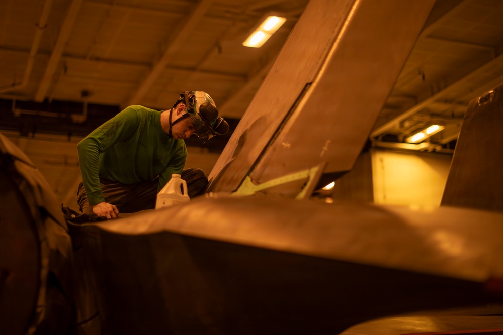 A technician wearing a green long-sleeve shirt and protective headgear is performing maintenance work on what appears to be a military aircraft in an enclosed hangar bay. The warm amber lighting suggests an indoor facility, likely a naval vessel's hangar deck. The worker is leaning over a large aircraft surface, possibly inspecting or servicing a component near the wing or fuselage area.