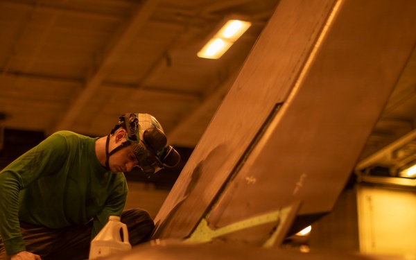 A technician wearing a green long-sleeve shirt and protective headgear is performing maintenance work on what appears to be a military aircraft in an enclosed hangar bay. The warm amber lighting suggests an indoor facility, likely a naval vessel's hangar deck. The worker is leaning over a large aircraft surface, possibly inspecting or servicing a component near the wing or fuselage area.