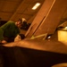 A technician wearing a green long-sleeve shirt and protective headgear is performing maintenance work on what appears to be a military aircraft in an enclosed hangar bay. The warm amber lighting suggests an indoor facility, likely a naval vessel's hangar deck. The worker is leaning over a large aircraft surface, possibly inspecting or servicing a component near the wing or fuselage area.