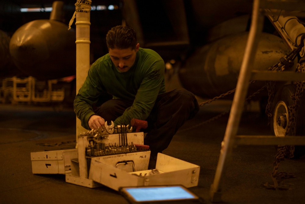 A person wearing a green long-sleeve shirt and dark pants is crouched down working with tools from an open equipment case or tool box. The setting appears to be a dimly lit industrial or military environment, possibly an aircraft hangar or ship deck, with large cylindrical objects and chained equipment visible in the background. A tablet device is visible on the floor in the foreground.
