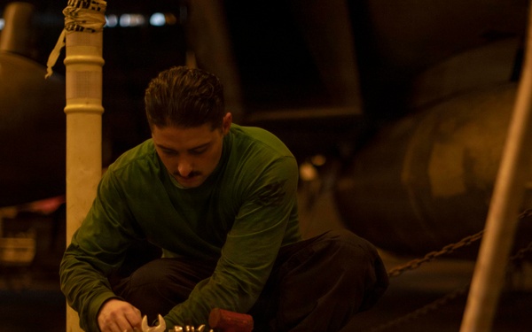 A person wearing a green long-sleeve shirt and dark pants is crouched down working with tools from an open equipment case or tool box. The setting appears to be a dimly lit industrial or military environment, possibly an aircraft hangar or ship deck, with large cylindrical objects and chained equipment visible in the background. A tablet device is visible on the floor in the foreground.