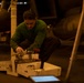 A person wearing a green long-sleeve shirt and dark pants is crouched down working with tools from an open equipment case or tool box. The setting appears to be a dimly lit industrial or military environment, possibly an aircraft hangar or ship deck, with large cylindrical objects and chained equipment visible in the background. A tablet device is visible on the floor in the foreground.