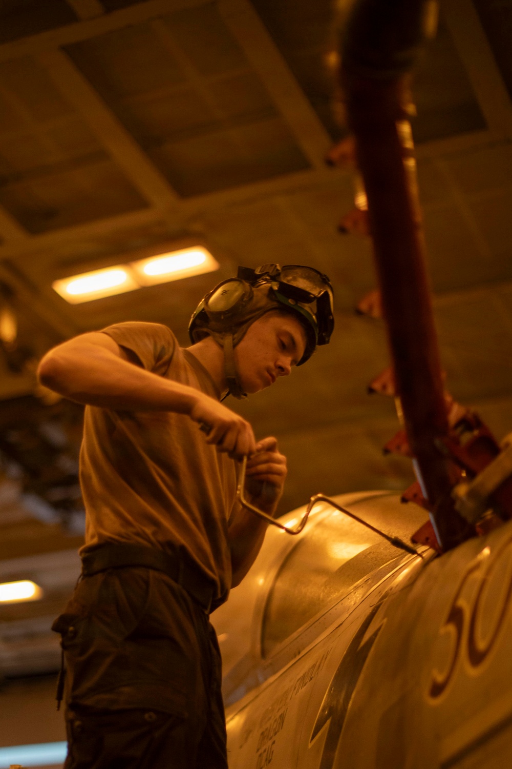 A young military technician or aviation mechanic is working on an aircraft in what appears to be a hangar bay, likely aboard a naval vessel. The individual is wearing a cranial helmet with goggles pushed up and is using a hand tool on the aircraft's fuselage. The warm amber lighting and enclosed overhead structure suggest an interior hangar deck environment.
