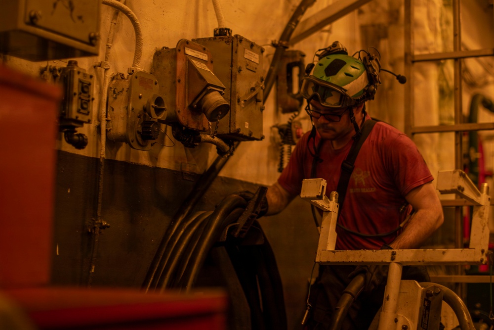 A worker in a red t-shirt and green safety helmet with attached communication headset and protective goggles is operating or inspecting heavy industrial equipment in what appears to be a confined industrial or mining environment. The scene is bathed in a warm orange/amber light, suggesting either artificial lighting conditions or smoke/dust in the atmosphere. The worker is interacting with large machinery mounted on a wall, which includes electrical junction boxes and heavy cables.