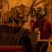 A worker in a red t-shirt and green safety helmet with attached communication headset and protective goggles is operating or inspecting heavy industrial equipment in what appears to be a confined industrial or mining environment. The scene is bathed in a warm orange/amber light, suggesting either artificial lighting conditions or smoke/dust in the atmosphere. The worker is interacting with large machinery mounted on a wall, which includes electrical junction boxes and heavy cables.