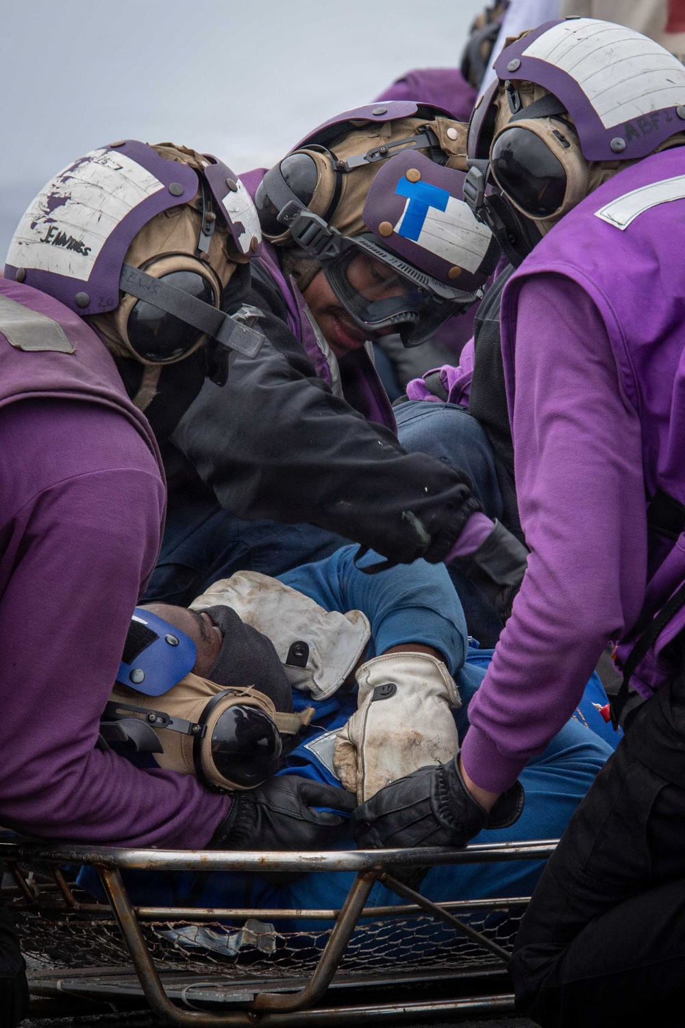 Several personnel wearing purple jerseys and flight deck helmets with goggles are attending to an injured or incapacitated person lying on a wire mesh Stokes litter/stretcher. The scene appears to be a casualty response drill or actual emergency on a naval aircraft carrier flight deck. The responders are wearing standard U.S. Navy aviation boatswain's mate (fuel) purple-colored flight deck gear.