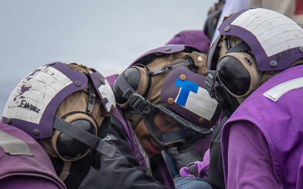 Several personnel wearing purple jerseys and flight deck helmets with goggles are attending to an injured or incapacitated person lying on a wire mesh Stokes litter/stretcher. The scene appears to be a casualty response drill or actual emergency on a naval aircraft carrier flight deck. The responders are wearing standard U.S. Navy aviation boatswain's mate (fuel) purple-colored flight deck gear.